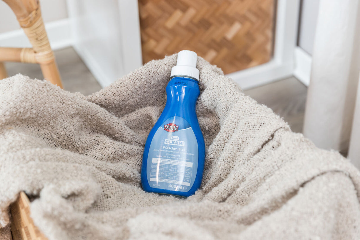 Eine blaue Flasche Waschmittel der Marke Trixie liegt auf einer beigen Decke in einem Korb.