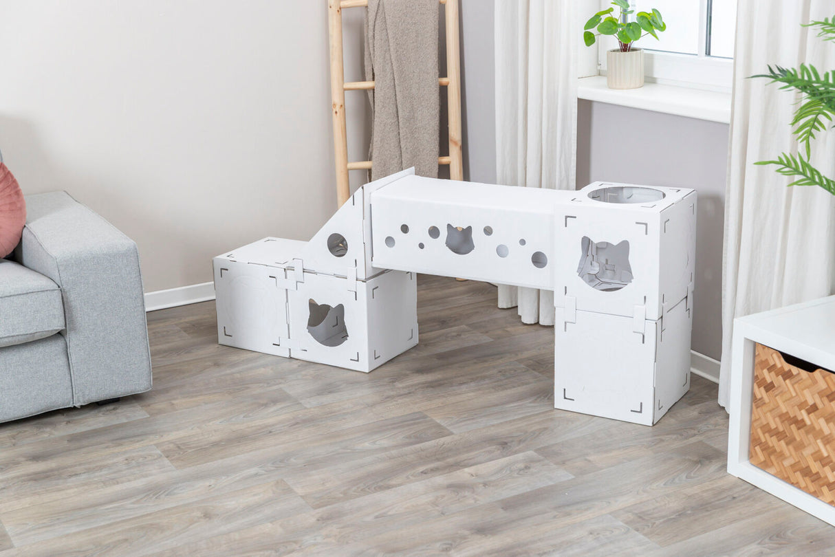 Weißes modulares Klettersystem mit Ausschnitten und Löchern, platziert auf hellem Holzfußboden neben grauem Sofa.