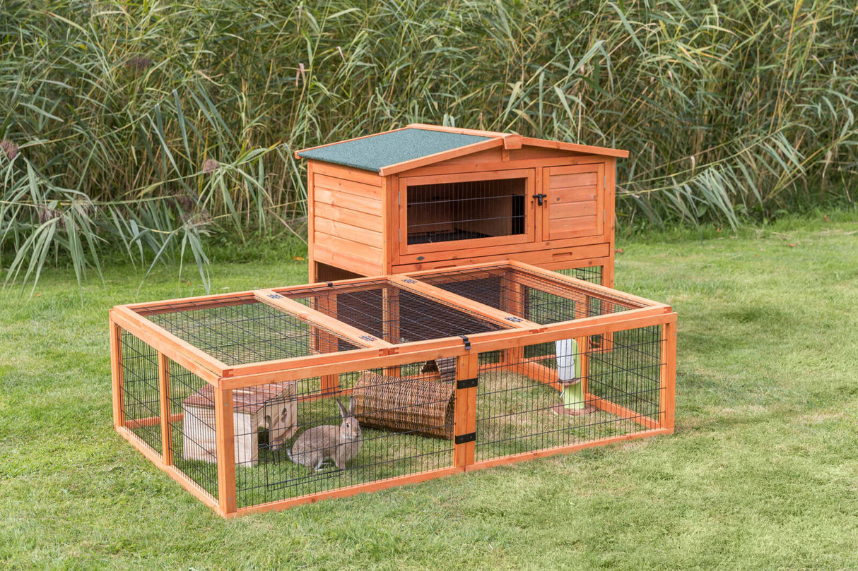 Holzkäfig auf einer Wiese mit zwei Kaninchen im Gehege, umgeben von hohem Gras, grünem Dach und Gitterabtrennung.