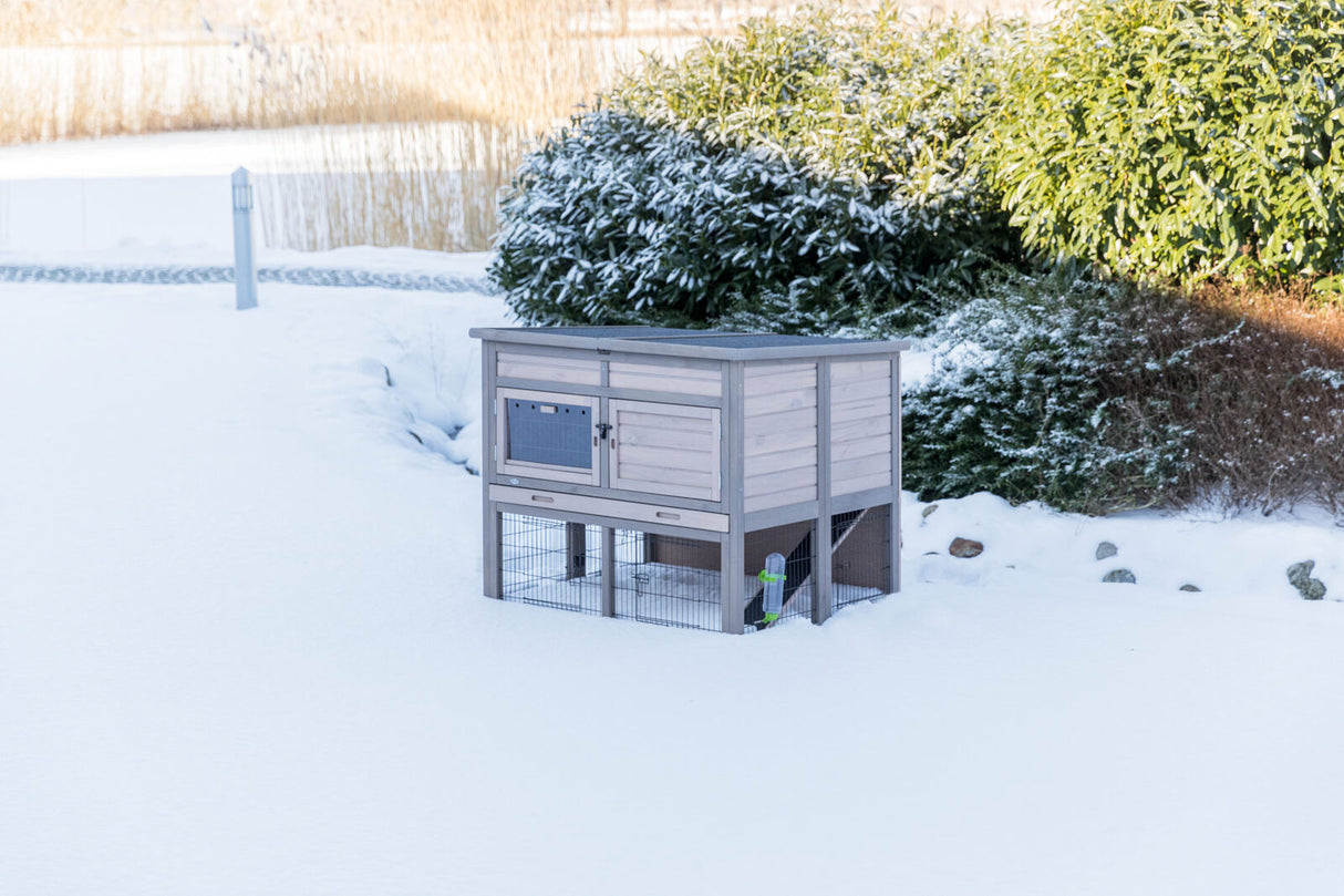 Ein grauer, hölzerner Außenstall mit Drahtgehege auf Schnee. Im Hintergrund grüne Büsche und Gräser.