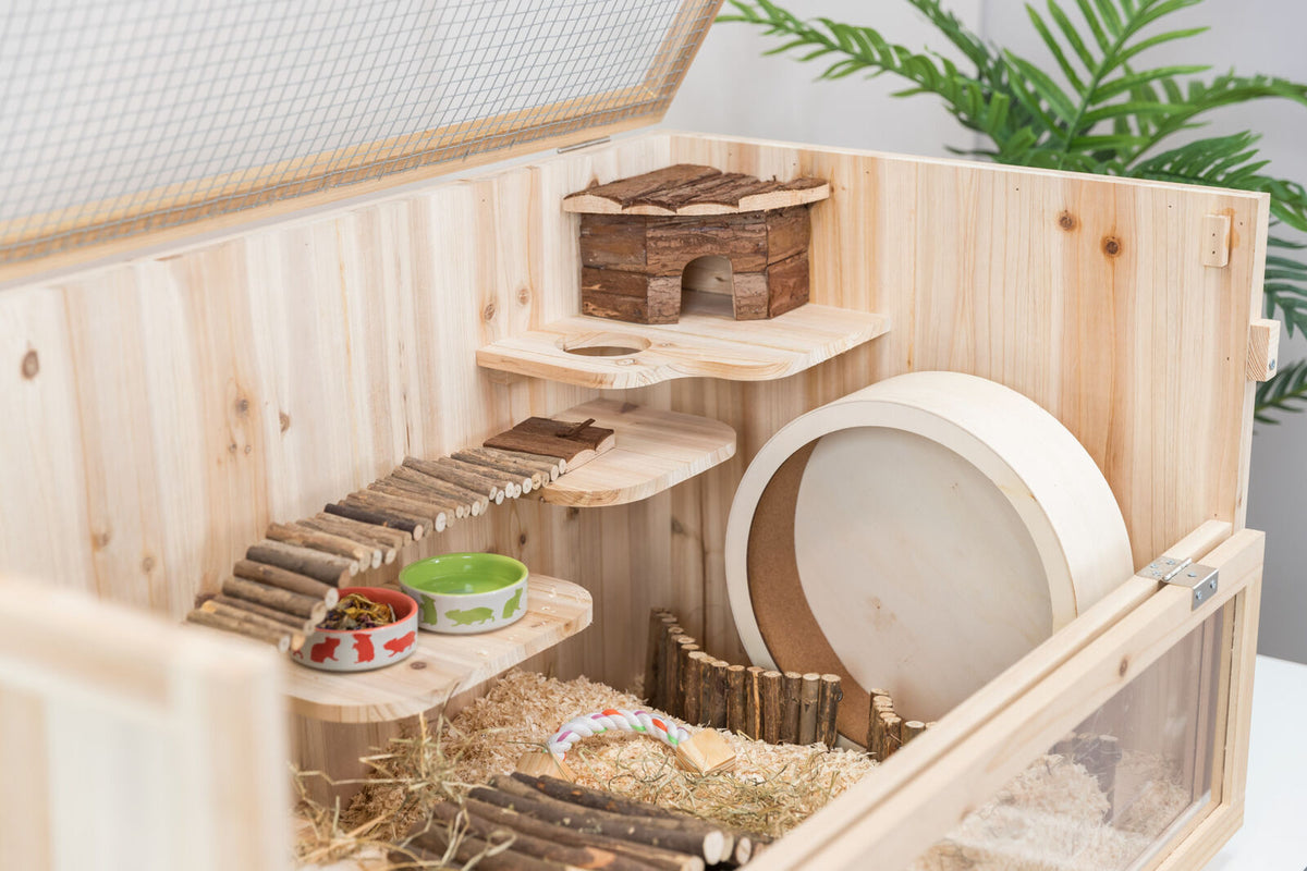 Holzkäfig mit Laufstall, Hütte, Treppen und Futterschalen, umgeben von Einstreu und Pflanzen im Hintergrund.