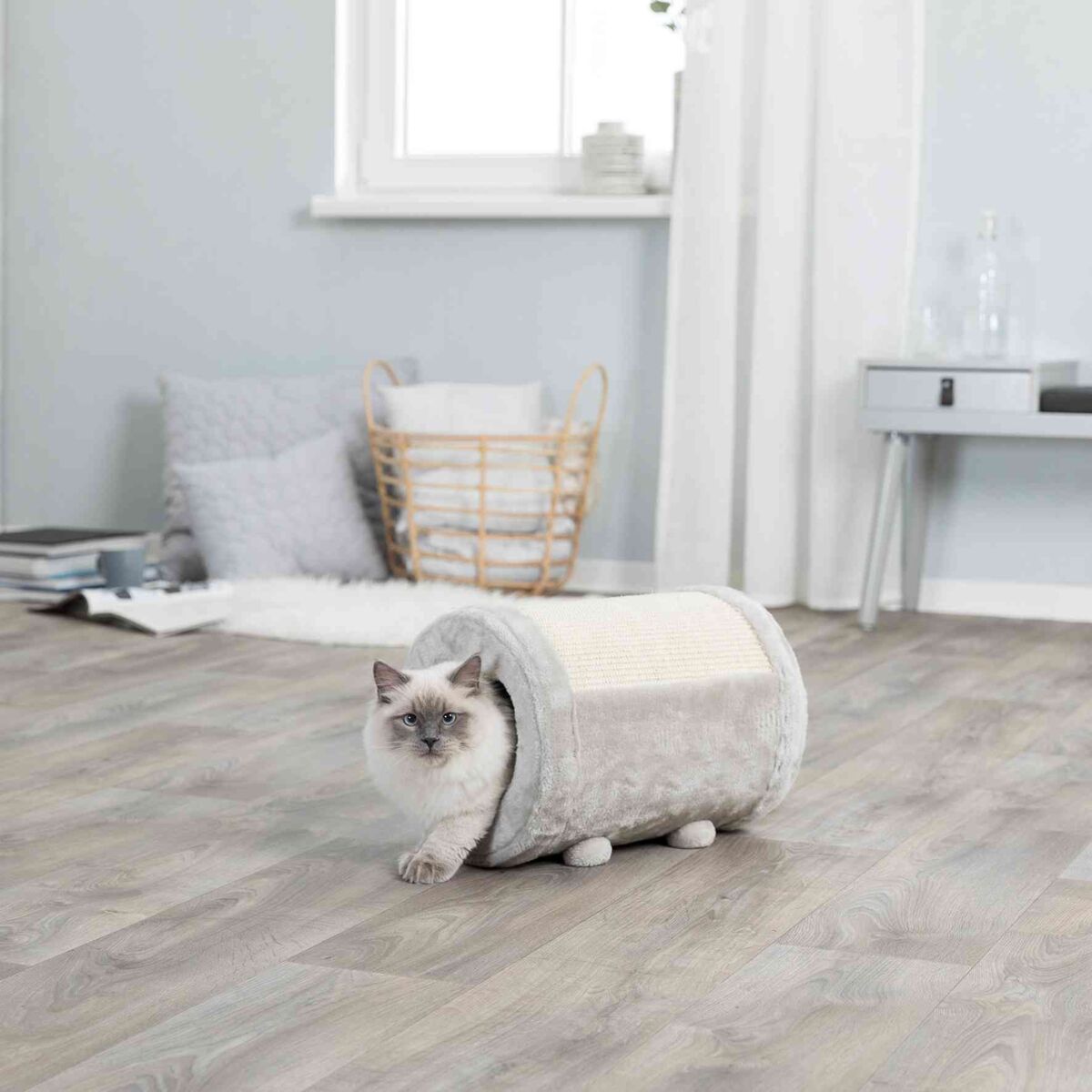 Eine graue Katze blickt aus einem pelzigen, zylindrischen Spielzeug in einem hellen Raum mit Holzboden heraus.