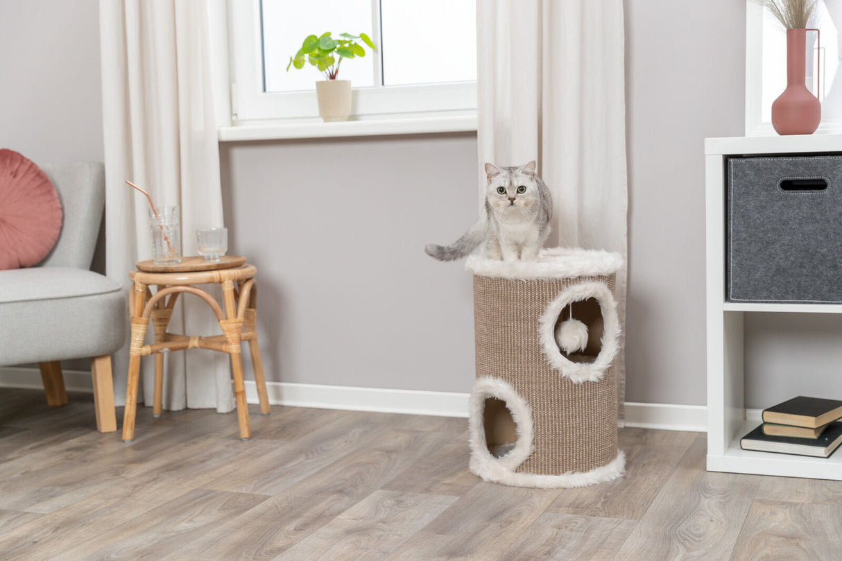 Eine graue Katze sitzt auf einem beige-weißen Kratzbaum in einem hellen Wohnzimmer mit Holzboden.