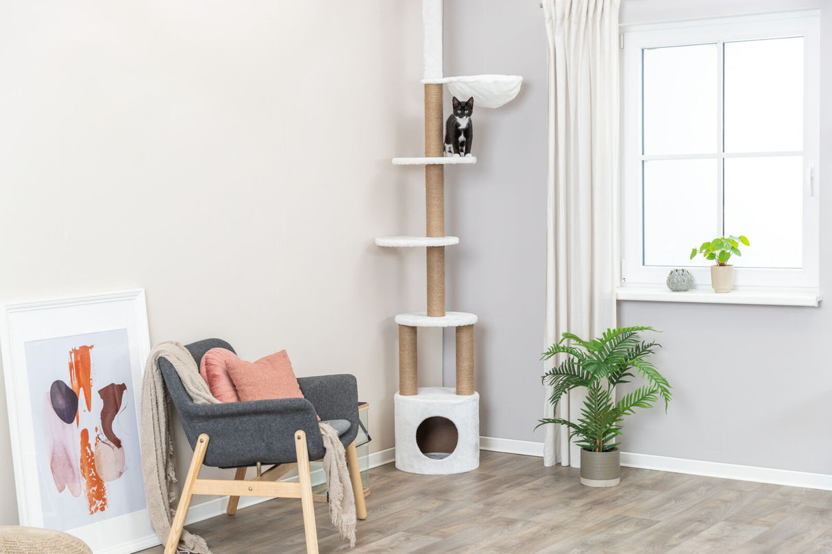 Wohnzimmer mit modernem Kratzbaum, Katze auf mittlerer Plattform, Sessel und Pflanzen, großes Fenster rechts.