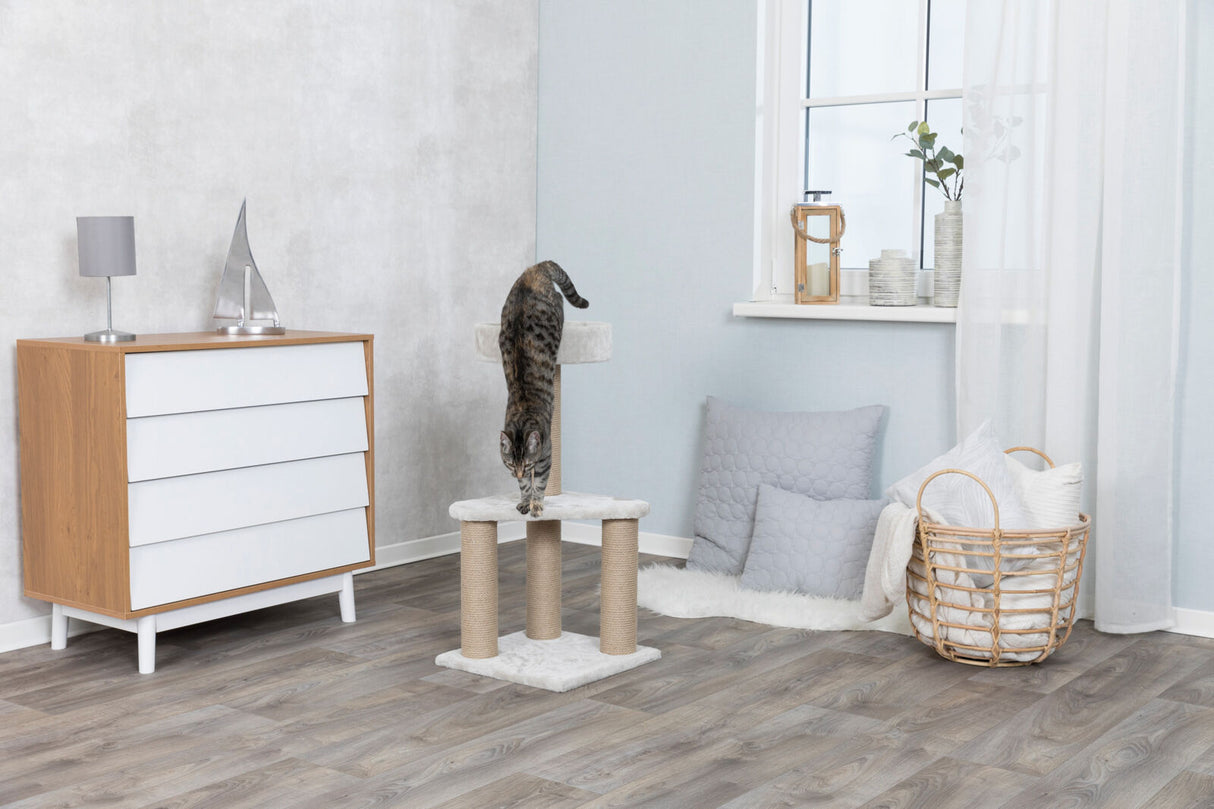 Katzenbaum mit Kratzsäulen in modernem Raum, daneben Holzkommode, Kissen, Korb und Fenster mit Deko.