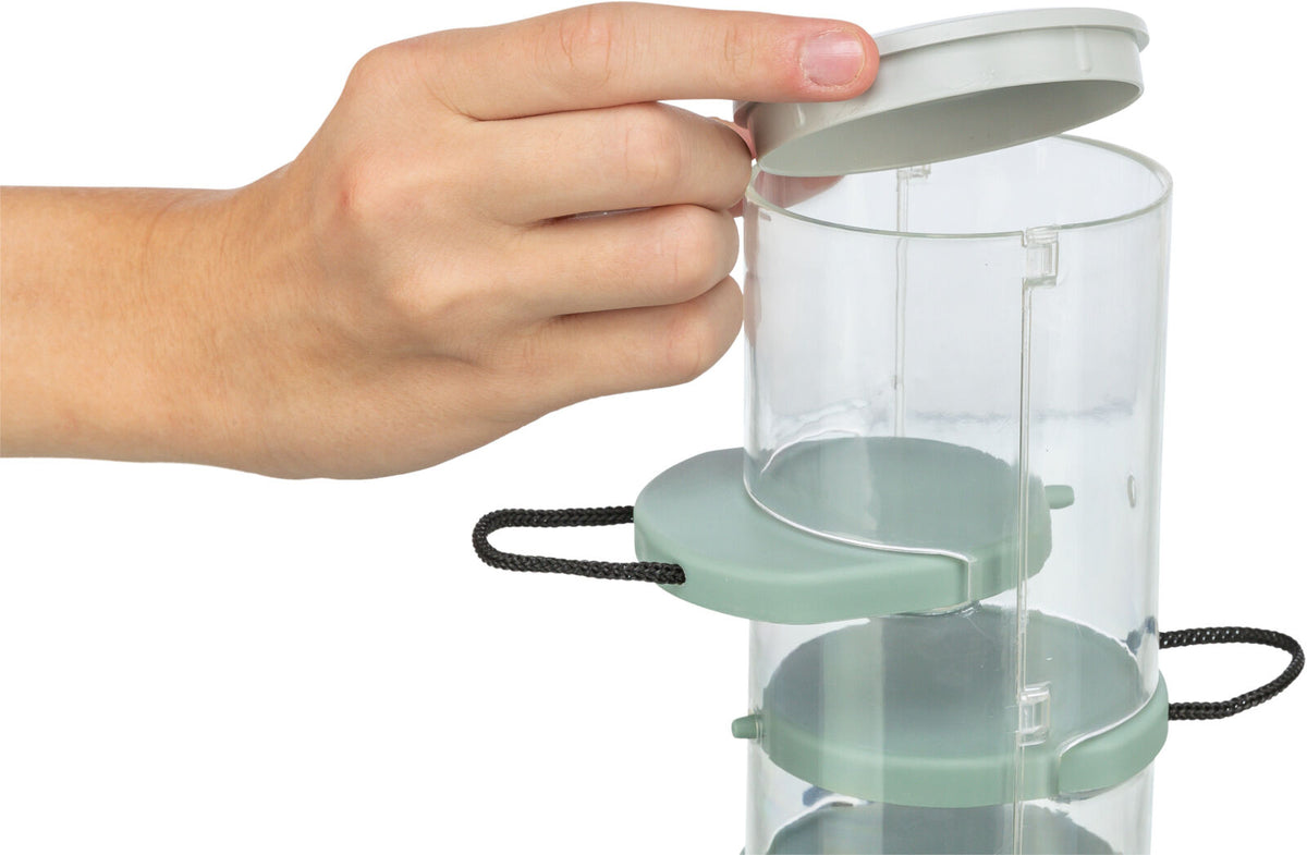 Hand öffnet einen transparenten, zylindrischen Behälter mit grünem Deckel und mehreren Ebenen, schwarzer Kordelgriff.