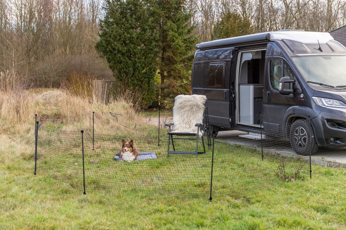 Wohnmobil mit offenem Türbereich steht neben einem eingezäunten Rasen, auf dem ein Hund auf einer Decke ruht.