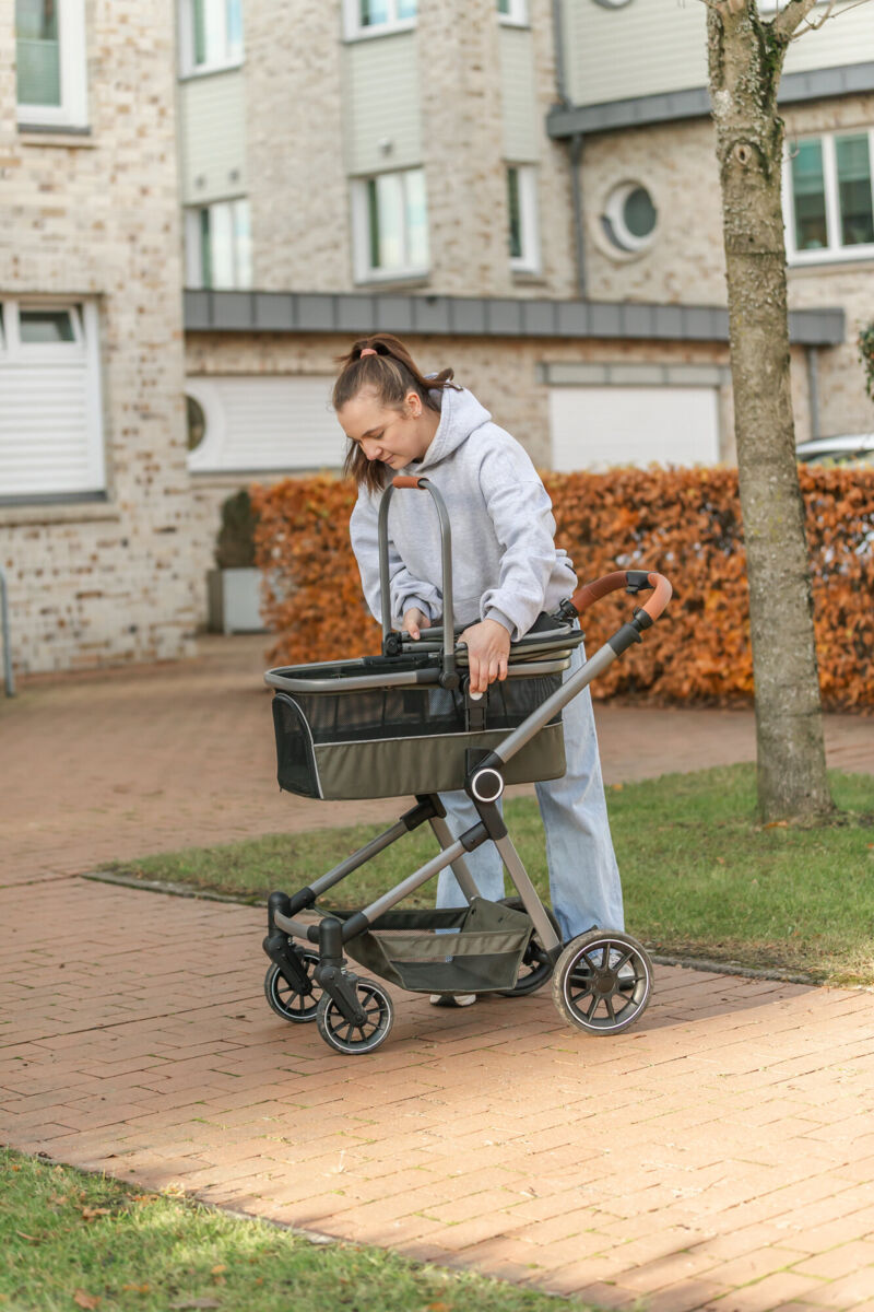 Person schiebt einen leeren Kinderwagen auf einem Gehweg vor einem Wohngebäude.