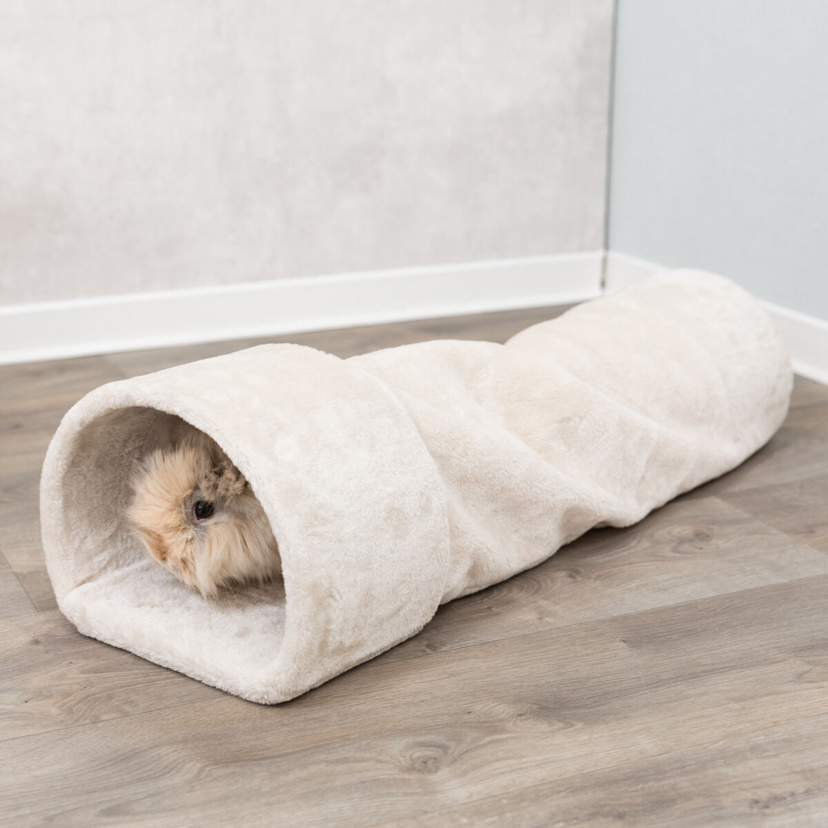 Flauschiger beige Tunnel liegt auf einem Holzboden, ein kleines pelziges Tier schaut heraus.