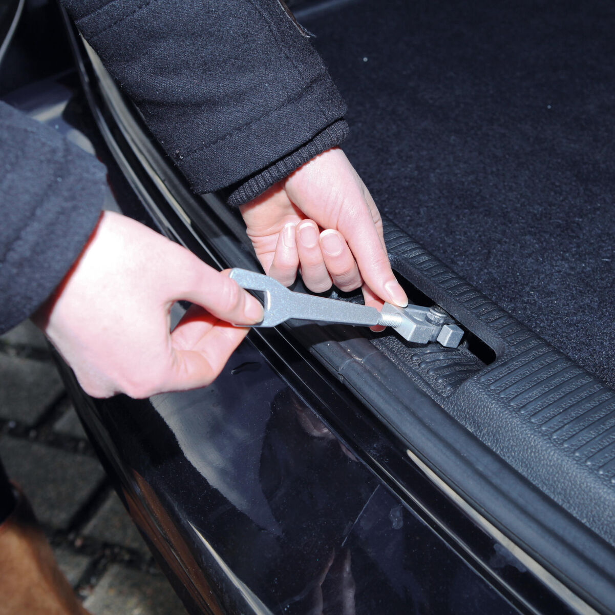 Eine Person hält ein metallisches Werkzeug und betätigt damit eine Schraube oder Mutter an einem schwarzen Kfz-Teil.