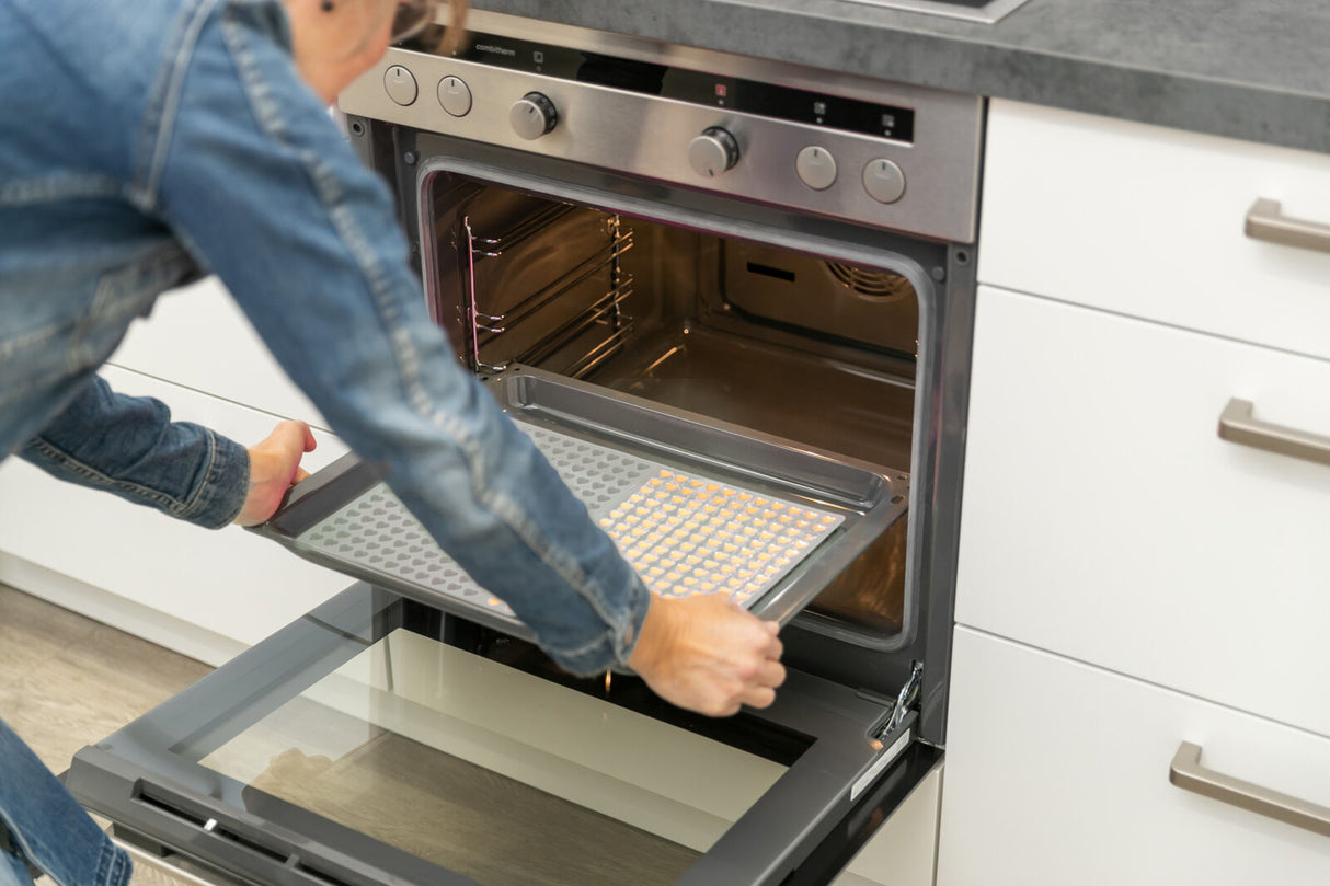 Eine Person schiebt ein Backblech mit kleinen gelben Objekten in einen offenen Backofen in einer modernen Küche.