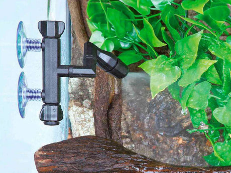 Schwarze Düse mit zwei Saugnäpfen an einer Terrarienscheibe, umgeben von grünen Blättern und Rinde.
