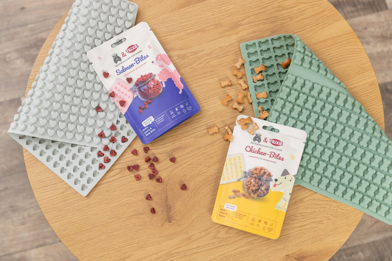 Backmatten in Mint und Grau mit verstreuten Leckerchen und zwei Packungen TRIXIE Snacks auf einem runden Holztisch