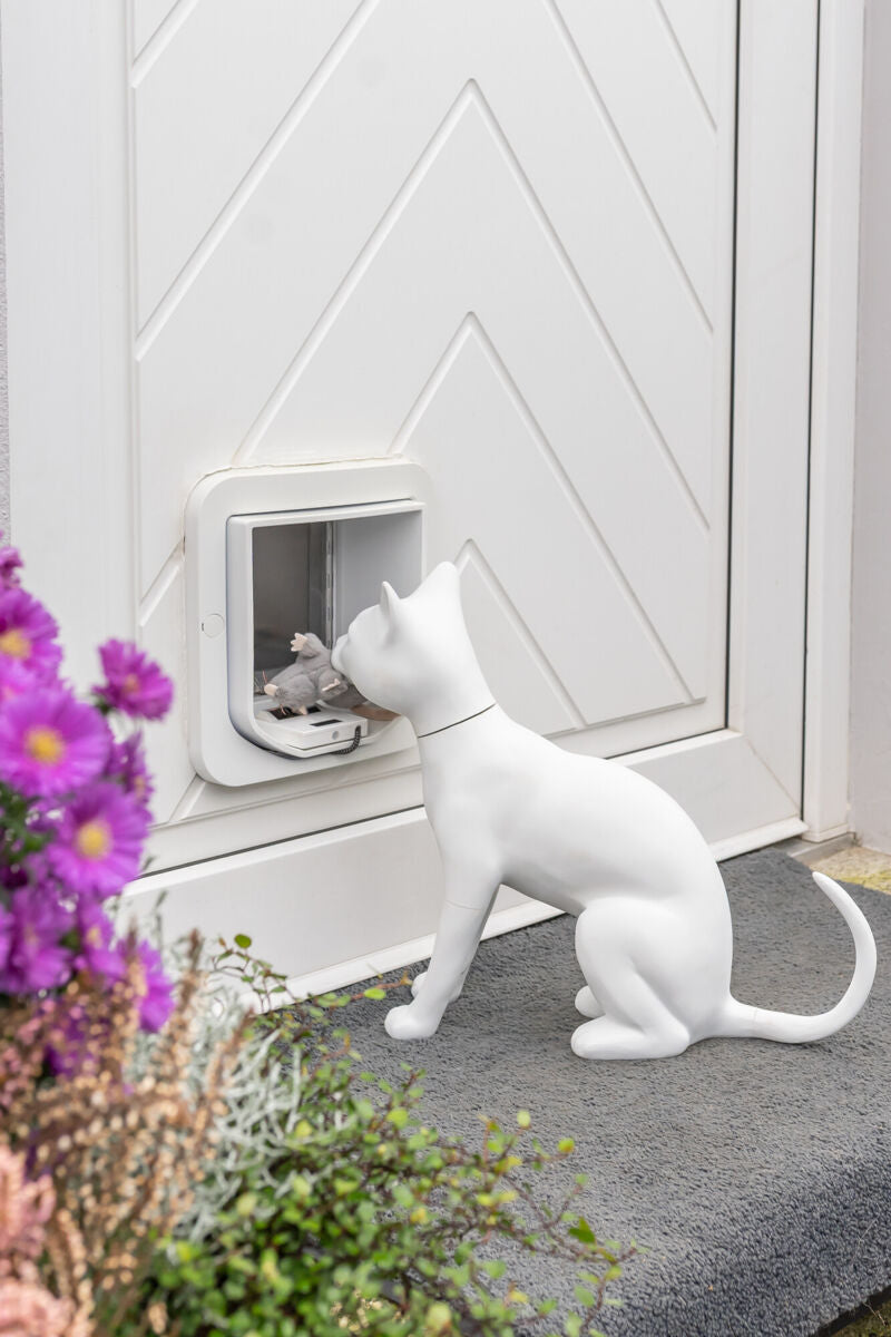 Weiße Katzenfigur vor Katzentür mit Spielzeugmaus, umgeben von Blumen.