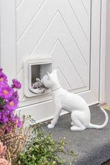 Weiße Katzenfigur vor Katzentür mit Spielzeugmaus, umgeben von Blumen.