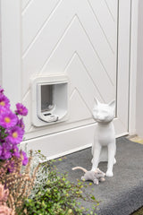 Weiße Katzenskulptur neben Katzenklappe auf grauem Teppich vor einer Tür.
