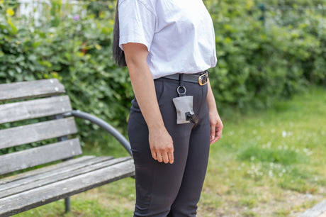 Eine Person trägt eine weiße Tüte an einem Gürtel, neben einer Holzbänke im Freien.