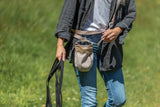 Person in Jeans und grauer Jacke hält schwarze Leinen und trägt eine kleine Tasche.
