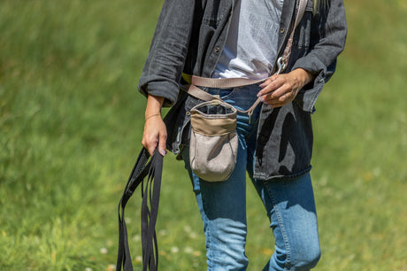 Person in Jeans und grauer Jacke hält schwarze Leinen und trägt eine kleine Tasche.