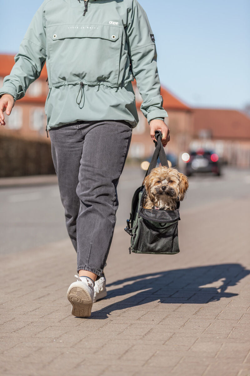 Person in grüner Jacke trägt einen Hund in einer schwarzen Tragetasche auf einem Gehweg.