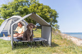 Ein brauner Hund sitzt in einem schattigen Hundebett vor einem Zelt am Strand.