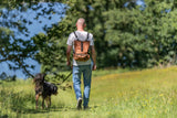 Mann mit braunem Rucksack geht mit Hund an der Leine auf einem grasbewachsenen Weg spazieren.