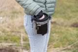 Person greift in eine kleine, graue Tasche mit schwarzem Netz auf einer Wiese.