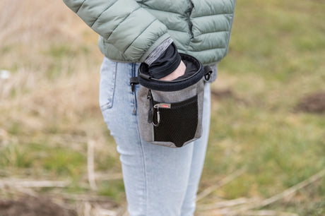 Person greift in eine kleine, graue Tasche mit schwarzem Netz auf einer Wiese.