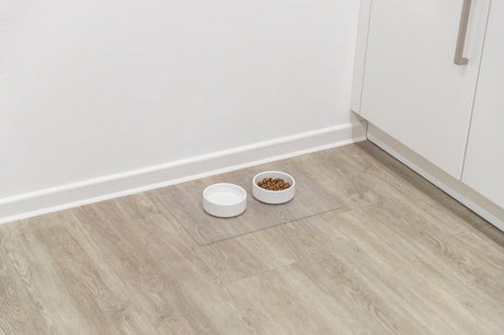 Zwei weiße Schalen auf einem Holzboden, eine mit Futter gefüllt, neben einer Küchenecke.