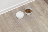 Zwei weiße Schüsseln auf einem Holzfußboden, eine mit Wasser gefüllt, die andere mit braunen Pellets.