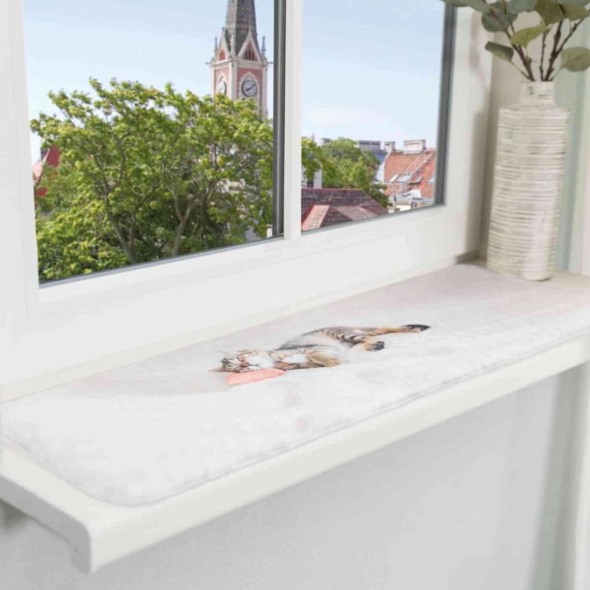Eine Katze liegt auf einem weißen Fensterbrett, daneben steht eine Vase, im Hintergrund ist ein Kirchturm zu sehen.