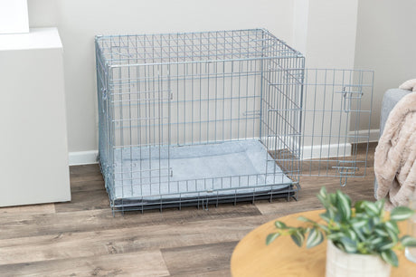 Offener Hundekäfig aus Metall mit grauer Matte steht auf Holzfußboden.