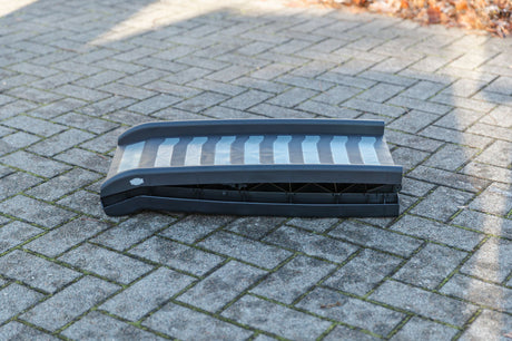 Eine schwarze, zusammenklappbare Rampe liegt auf einem gepflasterten Weg im Freien.