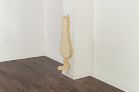 Ein beigefarbenes Kratzbrett in Form einer Katze hängt an einer weißen Wand über einem dunklen Holzboden.