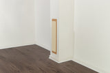 Katzenkratzbrett an einer weißen Wand, befestigt in einer Zimmerecke mit dunklem Holzfußboden.