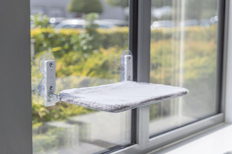 Katzenliege mit grauem Bezug ist an einem Fenster befestigt.