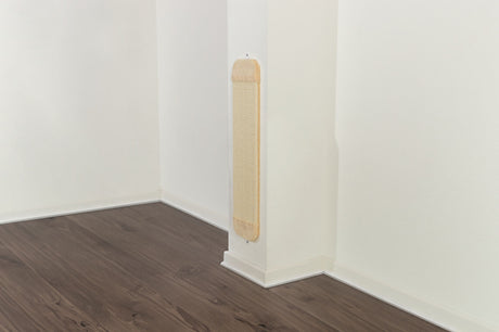 Kratzbrett mit beigem Rahmen an weißer Wand in einer Zimmerecke mit dunklem Holzboden.