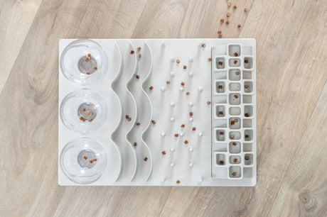 Weiße, wellenförmige Futterstation mit transparenten Schalen, verwendet für kleine braune Pellets auf hellem Holzboden.