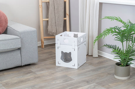 Weiße, würfelförmige Box mit ausgeschnittenem Katzenmotiv steht auf Holzboden neben grauem Sofa und Pflanze.