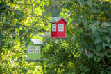 Zwei Vogelhäuschen in Hausform, eines rot und eines grün, hängen an Ästen in grünem Laubwerk.