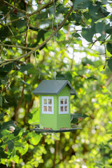 Grünes Vogelhaus in Form eines Häuschens, gefüllt mit Samen, hängt an einem Ast vor grünem Blätterhintergrund.
