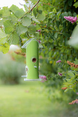 Ein grüner Vogelfutterspender hängt an einem Ast mit dichtem Laub und Blumen im Hintergrund.