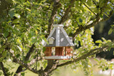 Ein kleines, hölzernes Vogelhaus mit grauem Dach und weißem Rahmen hängt an einem grünen Baum.