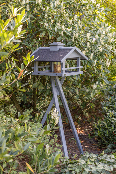 Ein graues Vogelhäuschen mit schwarzem Dach steht auf einem dreibeinigen Ständer vor dichtem grünem Laub.