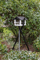 Ein weiß-schwarzes Vogelhaus auf einem dreibeinigen Ständer steht vor einem dichten grünen Busch.