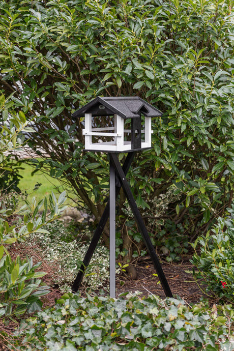 Ein weiß-schwarzes Vogelhaus auf einem dreibeinigen Ständer steht vor einem dichten grünen Busch.