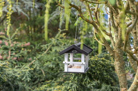 Vogelfutterhaus aus Holz mit schwarzem Dach, aufgehängt an einem Baum in einem grünen Gartenumfeld.