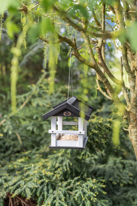 Weiße und schwarze Vogelhäuschen aus Holz hängt an einem Baum, umgeben von grünen Blättern und Zweigen.
