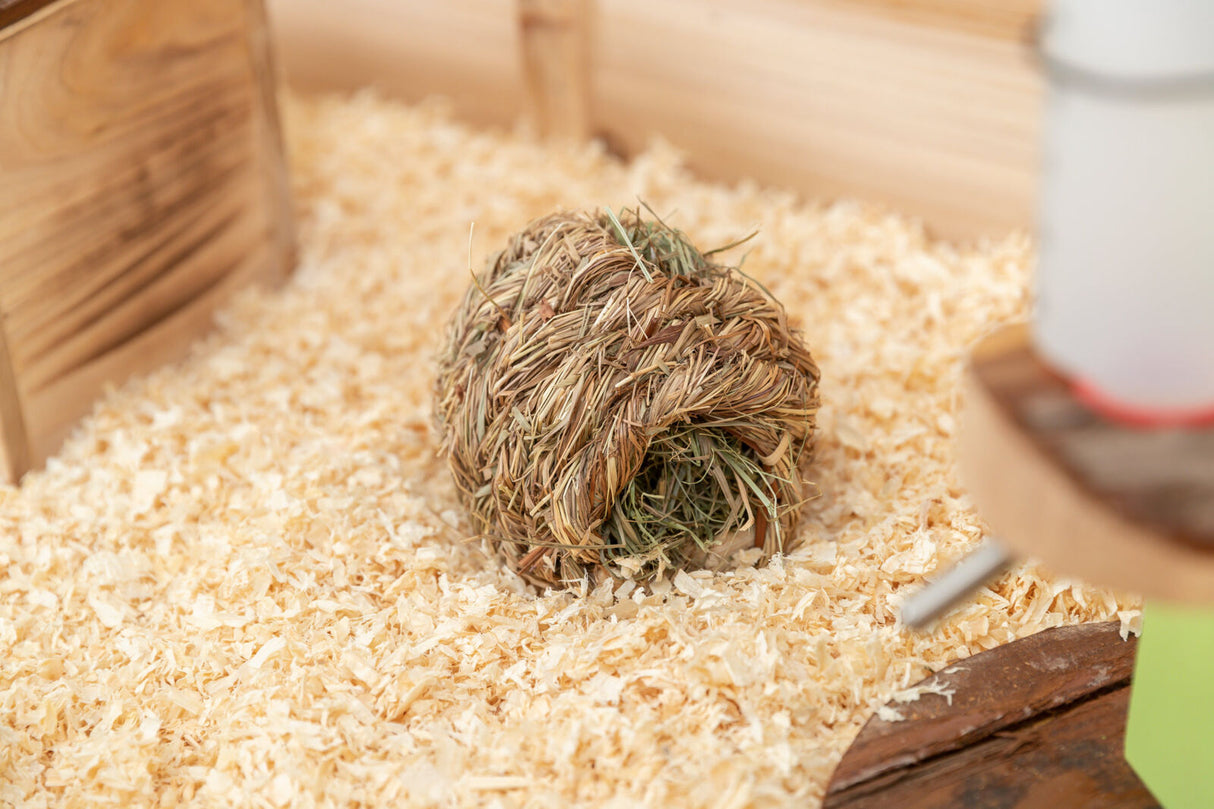 Geflochtenes Ballspielzeug aus Stroh auf Holzspänen vor Holzwänden und einem unscharfen Trinkspender.