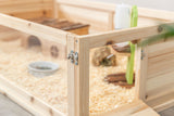 Holzgehege mit Transparentwand, innen Holzspäne, eine kleine Hütte, Futternapf und ein Spielzeug sichtbar.
